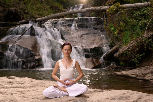 Young woman sitting in lotus pose under tropical waterfall meditating with calm strength and mindfulness fully connected to nature energy tranquility and inner balance - Powered by Adobe