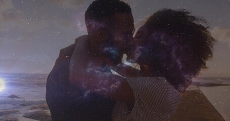 Embracing couple wearing striped shirt kissing on wooden pier at dusk, waves, nebula overlay