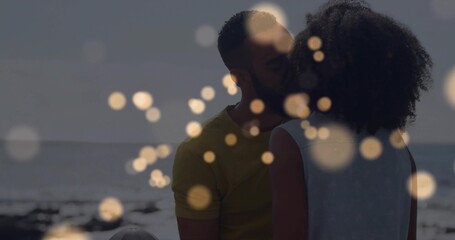 Kissing couple wearing summer attire on sandy shore, with ocean waves horizon line and bokeh lights