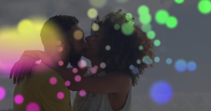 Kissing Black woman and bicultural man embracing at calm shoreline under clear sky, colorful bokeh