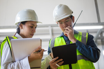Two engineers in safety gear collaborate on industrial project using tablet and documents. They are focused and engaged in modern industrial setting