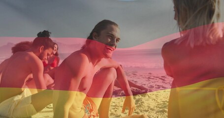Smiling group of friends in swimsuits sitting on sandy shore at water's edge, with warm sunlight