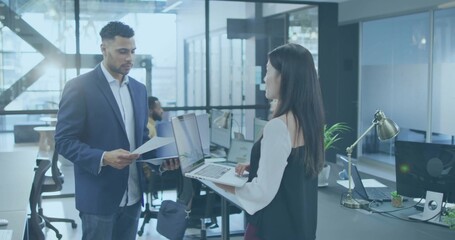 Colleagues wearing business attire reviewing documents and sharing laptop in office, with monitors