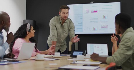 Presenting mid adult man leaning on conference table in modern office with large chart display