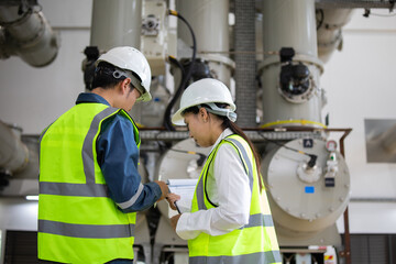 Two engineers in safety gear collaborate in industrial setting, discussing plans in front of large machinery. Their teamwork highlights importance of safety and precision