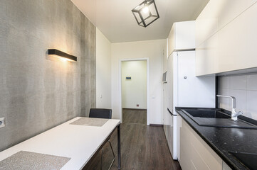 modern kitchen with white cabinets and a black countertop. A white table sits near a textured wall with a black light fixture. A doorway leads to another room