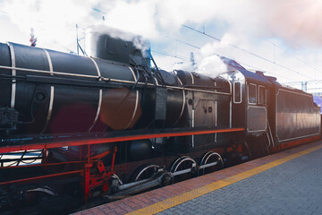 A vintage black steam locomotive emitting smoke stands at a railway station platform under a cloudy sky