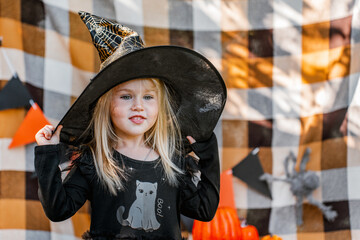 A little girl in a black dress and a witch hat dressed up for Halloween, flags on an orange plaid...