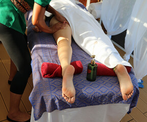 Asian masseuse giving a vigorous massage to a reclining womans leg on a massage table during a spa...