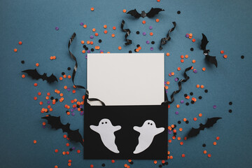 Halloween party composition with empty card decorated with bats, ghosts and confetti on dark blue background top view.