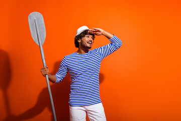 Positive man in striped sailor-inspired shirt holding a paddle posing cheerfully against an orange...