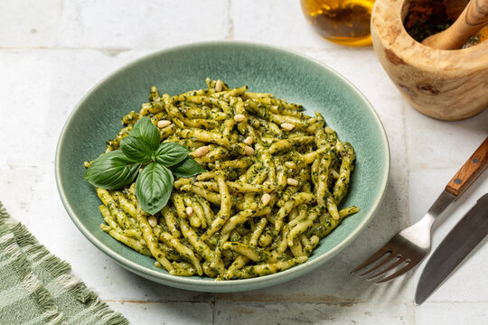 Cloae up of a plate with Trofie al pesto,  italian pasta with homemade pesto genovese, made with basil leaves, olive oil, parmesan, pecorino, pine nuts, and garlic. White table surface.
