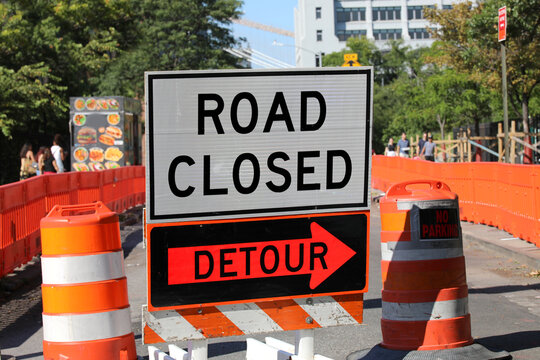 road closed sign with a detour arrow and orange construction barrels on a street