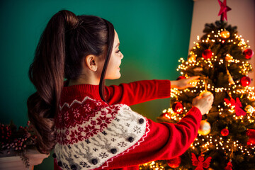 Festive young woman decorates a beautifully lit Christmas tree at home, spreading holiday cheer and...