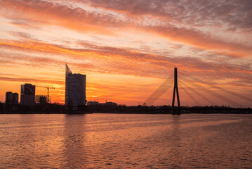 Fototapeta premium Photograph of a sunset view over a river with a bridge and a high-rise building in the background. The scene is bathed in warm orange tones, creating a dramatic and atmospheric cityscape.