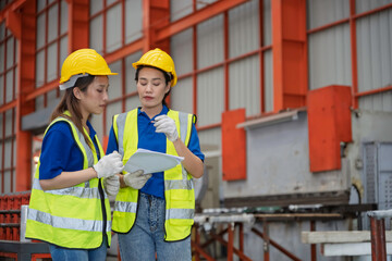 Two female factory workers wearing safety helmets and vests discussing work tasks in a modern industrial plant. Concept of female engineers, industrial teamwork, and workplace safety.