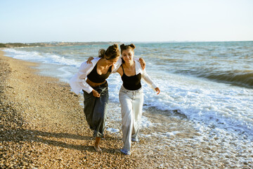 Two teenage girls 15-16 year old wearing casual clothes denim pants and white shirts having fun at beach over sea outdoors. Summer vacation season. Teenagerhood.