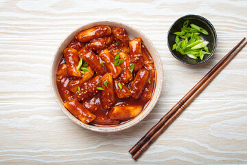 Stir fried rice cakes tteokbokki simmered in spicy sauce, garnished with green onions and sesame seeds, served on a white wooden background with chopsticks top view. Popular dish of Korea