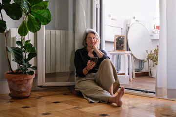 Serene mature woman enjoying quiet moment at home, browsing smartphone while sitting on the floor near balcony, embracing peaceful atmosphere