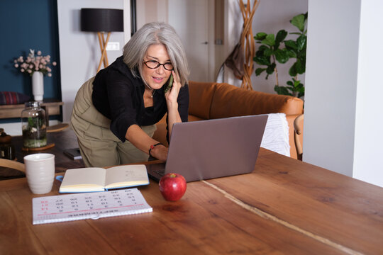 Confident mature businesswoman talking on phone and working on laptop at home office