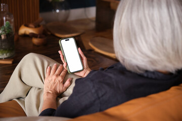 Elderly woman holding smartphone with white screen relaxing on couch, over shoulder view from behind