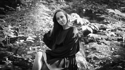 A young woman in a black dress standing barefoot in the forest, leaning against a tree, surrounded...