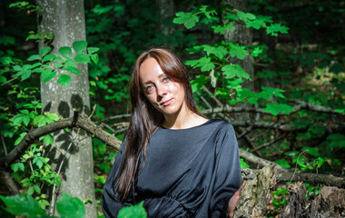 A young woman in a black dress standing barefoot in the forest, leaning against a tree, surrounded by green leaves and natural sunlight. Concept of harmony with nature, freedom, and natural beauty.