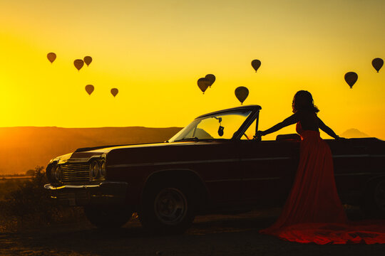 Silhouette of a Woman in Red Dress and Vintage Convertible Car at Sunrise with Hot Air Balloons in Cappadocia