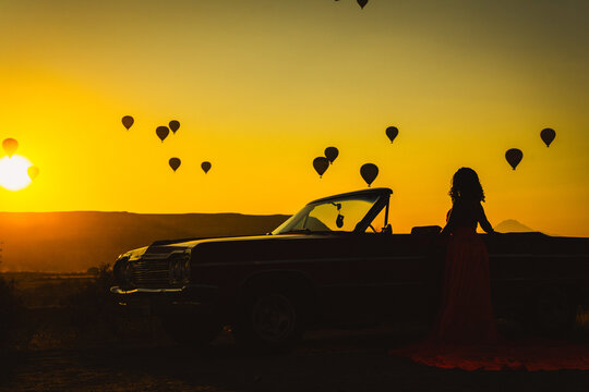 Silhouette of a Woman in Red Dress and Vintage Convertible Car at Sunrise with Hot Air Balloons in Cappadocia