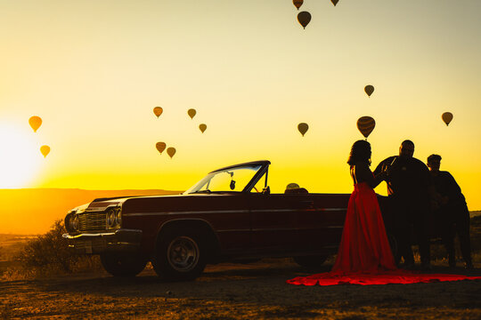 Silhouette of a Woman in Red Dress and Vintage Convertible Car at Sunrise with Hot Air Balloons in Cappadocia