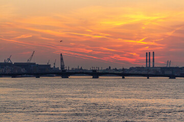Obraz premium Panoramic sunset view from Palace Bridge showing Neva River with orange sky reflection, Blagoveshchenskiy Bridge and industrial port cranes. Saint Petersburg, Russia.