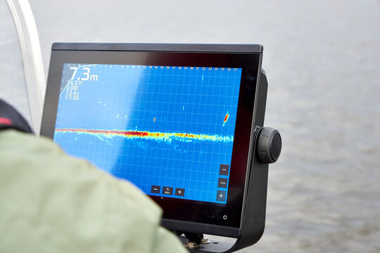 Fisherman steering boat while looking at fishfinder screen on a lake. Modern angling technology with echo sounder and scenic river view. Fishing, navigation, and outdoor recreation concept