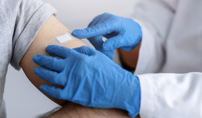 Remote medicine, clinic services and patient health care at home. Doctor in white coat and protective gloves puts band aid to shoulder of arab young man with beard in mask on gray background, cropped