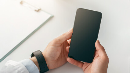 Workplace, home office, business, medical website and blog. Millennial doctor in coat with smart watch uses smartphone with blank screen on white desk with notepads and glass of water, free space