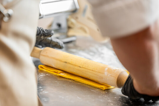 Chef in black gloves rolling yellow dough on floured surface with pasta machine in background. Professional kitchen scene, ideal for culinary, baking, or pasta-making themes. - Powered by Adobe