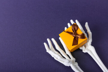 Halloween skeleton hands holding spooky gift box on purple background, copy space