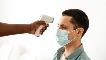 Health protection, immunization procedure and prepare for vaccination in clinic. African american doctor use forehead thermometer to check temperature for virus covid-19 symptoms to man in mask