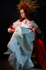 Fototapeta premium A young woman dressed in a national costume with a red wreath and a headgear made of wheat is sitting on a chair, casting a mysterious and elegant look in dim lighting.