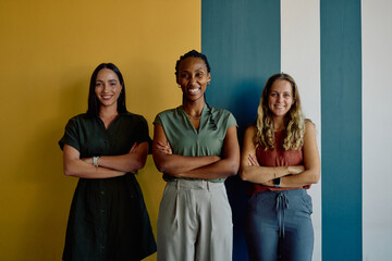 Portrait of smiling diverse businesswomen standing arms crossed against colorful wall in office