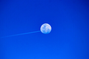 Moon, plane, and contrail against a brilliant blue sky