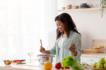 Pretty housewife tasting fresh meal at home. Chef taste food, cook, covid-19. Smiling young african american lady make soup in modern kitchen interior with bright vegetables, on window background