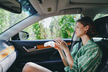 Woman seated in car interior, wiping lips with a tissue, casual striped shirt, daytime light and...