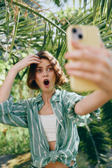 Young woman in a green striped shirt takes a surprised selfie in a lush garden, sunlight filtering...