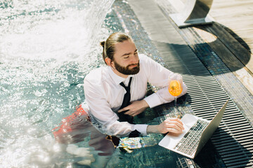 Businessman working on laptop in swimming pool with cocktail