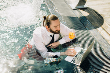 Businessman working on laptop in swimming pool with cocktail