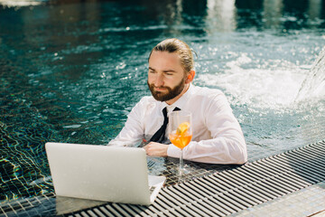 Businessman working on laptop in swimming pool with cocktail