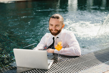 Businessman working on laptop and drinking cocktail in swimming pool