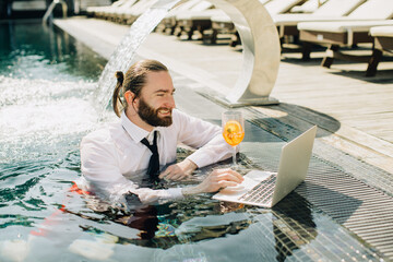 Businessman working on laptop in swimming pool with cocktail