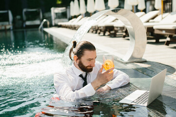 Businessman working remotely from swimming pool with cocktail and laptop