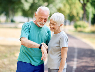 Smiling active senior couple jogging exercising and having fun and laughing together taking a break in the park, senior athletes checking their er heart rate with smartwatch